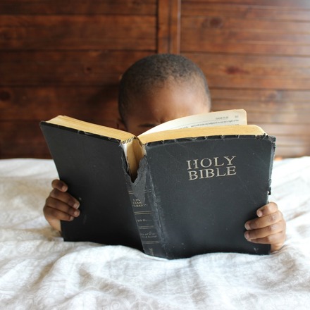 African child reading tattered bible on white bed